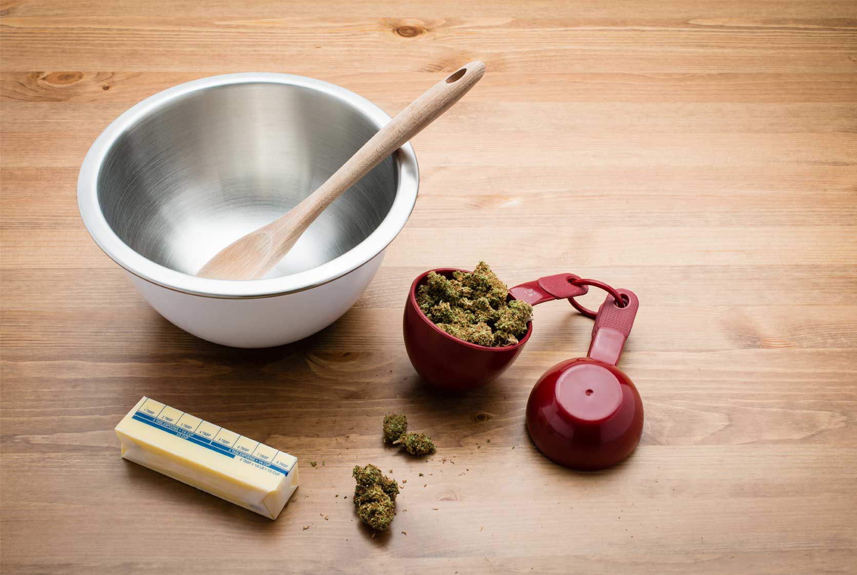 red measuring cup with cannabis flower next to a stick of butter and a metal mixing bowl with a wood spoon on a table