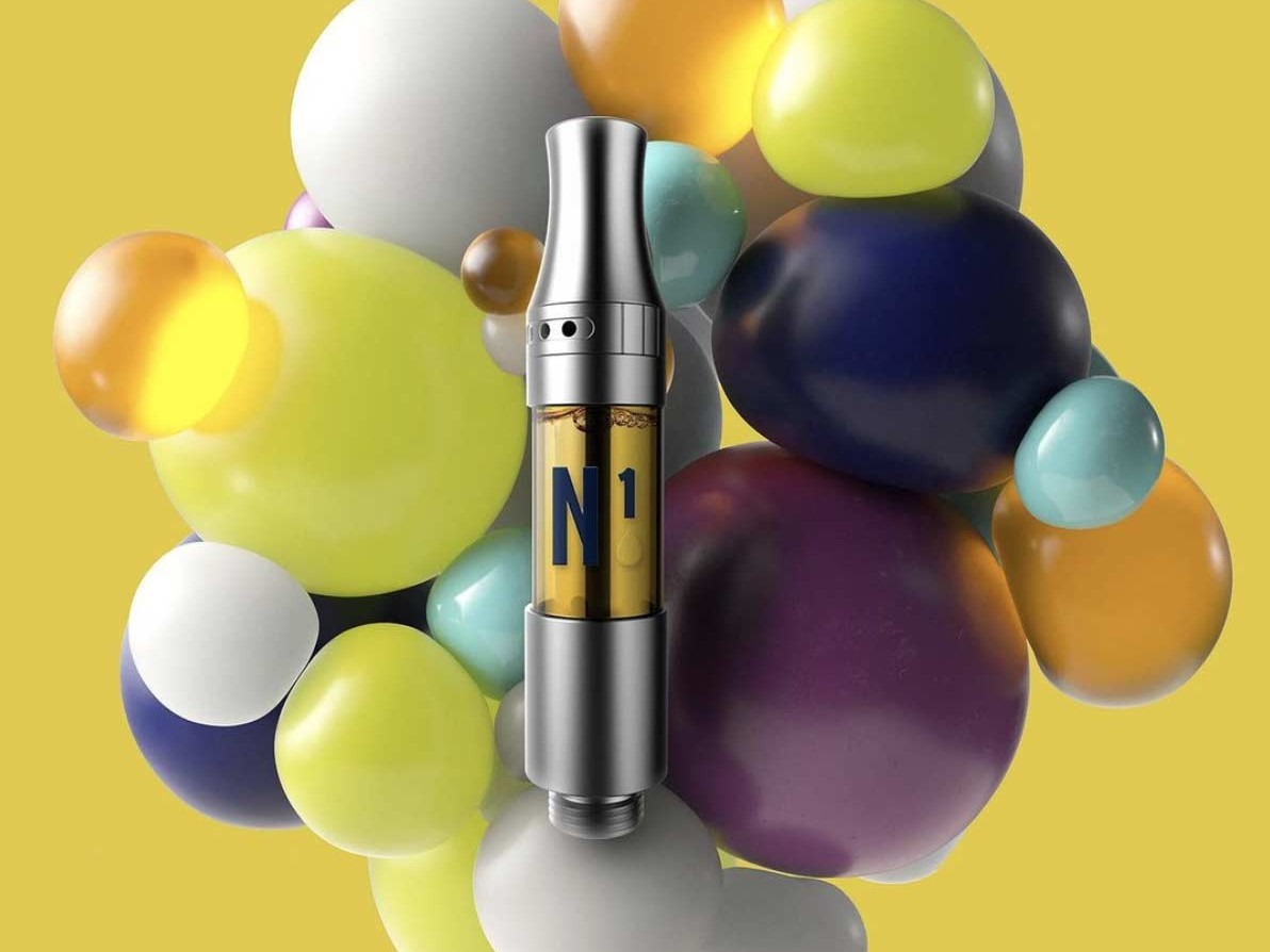 A Next 1 cannabis vape cartridge filled with golden oil, featuring a sleek metallic design, positioned in front of a vibrant arrangement of multicolored spheres on a yellow background.