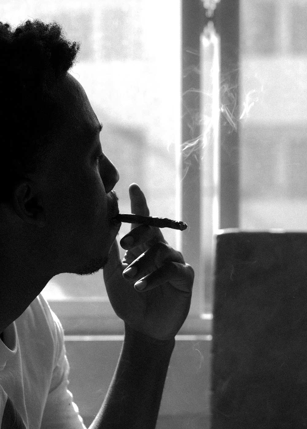 grayscale picture of a man smoking a joint looking out a window